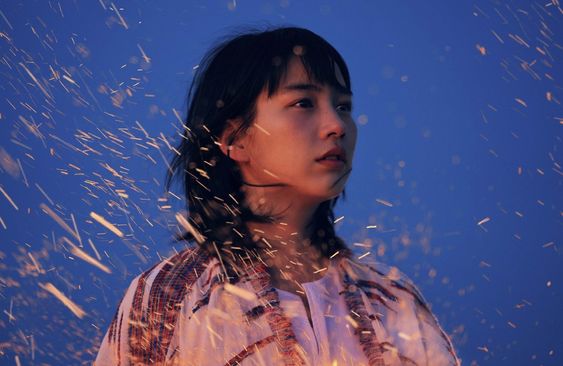 a young woman with dark hair stands against a backdrop of a twilight blue sky filled with cascading sparks, possibly from a bonfire or fireworks. she wears a lightcolored, patterned top and her expression is contemplative, her gaze directed off to the side. the closeup perspective emphasizes her features and the dramatic lighting creates a sense of intensity and wonder.