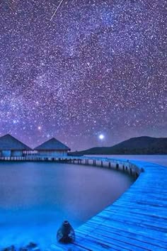 a serene landscape with a wooden bridge extending across a still body of water.  two huts sit in the distance, partially submerged, creating a sense of tranquility. the sky above is ablaze with stars, forming a breathtaking display of the milky way galaxy, adding to the magical atmosphere of the scene.