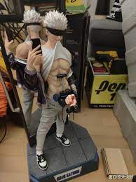 loseup view of a collectible figurine, possibly from an anime or manga, depicting a male character with spiky white hair, wearing a gray hoodie and sneakers. the figurine is posed as if taking a selfie with a phone and holding a game controller. it's displayed on a dark gray base and is partially reflected in a mirror behind it.  the background shows various boxes and packaging, suggesting it is in a retail or storage setting.