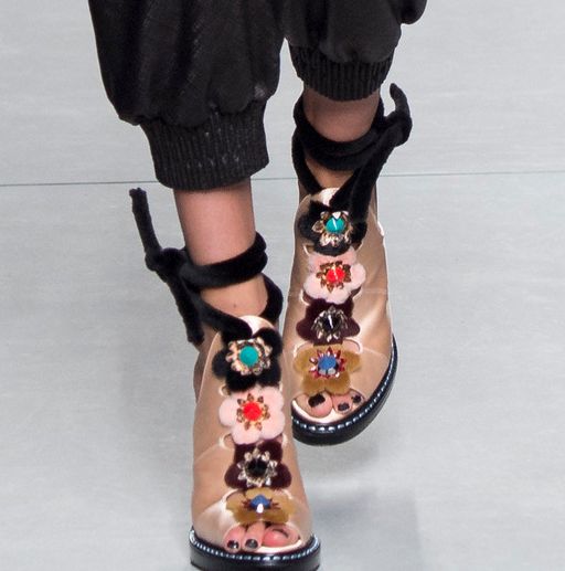 this image shows a model wearing a pair of decorative sandals. the sandals are made of tancolored material and have a platform heel, with black straps and multiple jeweled flower decorations. the model is wearing black pants and has their legs crossed in the image.