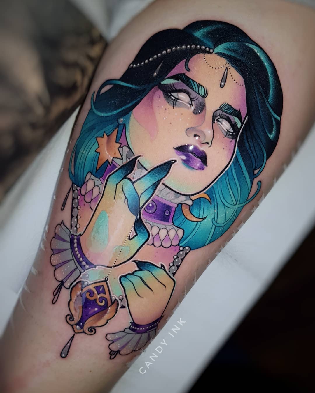 this is a neotraditional style tattoo on someone's upper arm of a woman with blue hair, purple lipstick, and mystical details like a sun and moon in her hair. her fingers are elongated, and she is touching her chin. there are also purple crystals, pearls, and jewels. the background of the tattoo is unshaded skin.