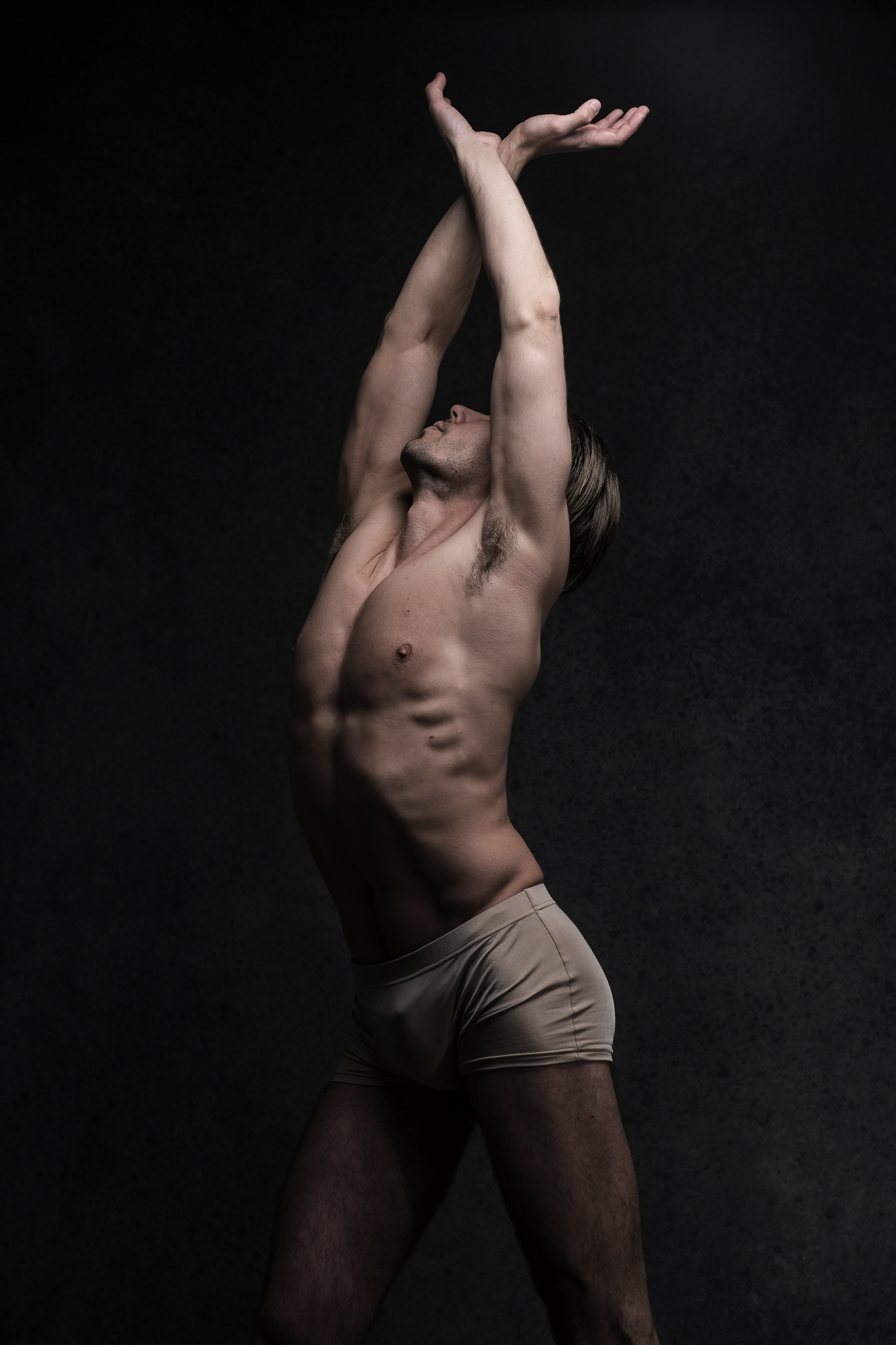 a shirtless man with toned muscles is standing against a dark gray background and stretching his arms above his head with his eyes closed. he is wearing beige briefs. the image is lit with spotlights, highlighting his physique and creating a dramatic effect.