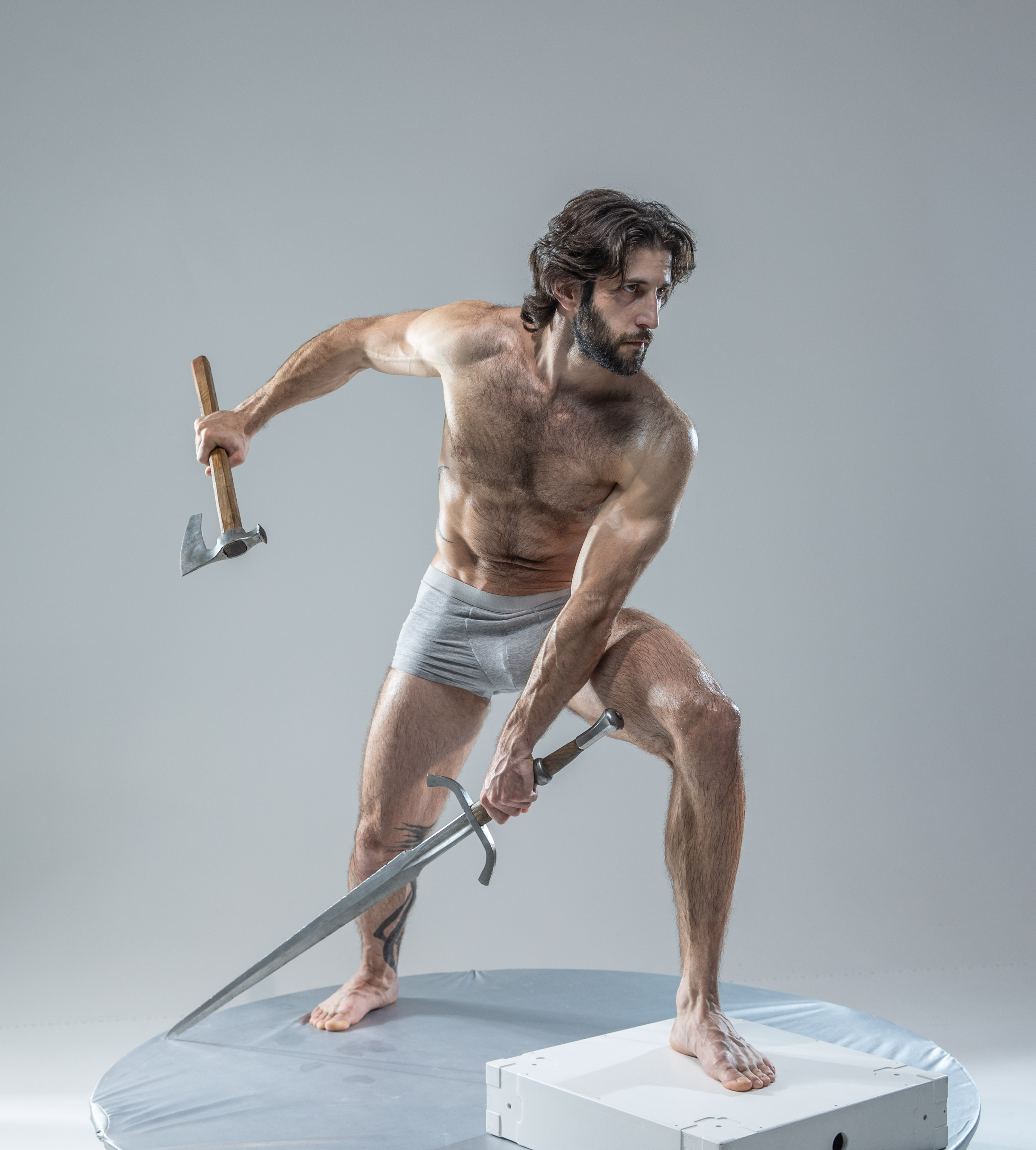 a muscular man with long hair and beard, wearing only underwear, stands on a round platform in a fighting stance. he holds a sword in his right hand and an axe in his left hand. he's illuminated by soft lighting against a plain grey background, creating a dramatic and powerful image.
