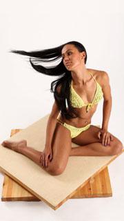 a woman with long black hair is posing on a wooden platform. she is wearing a yellow bikini and is looking to the right of the frame.  the background is white.