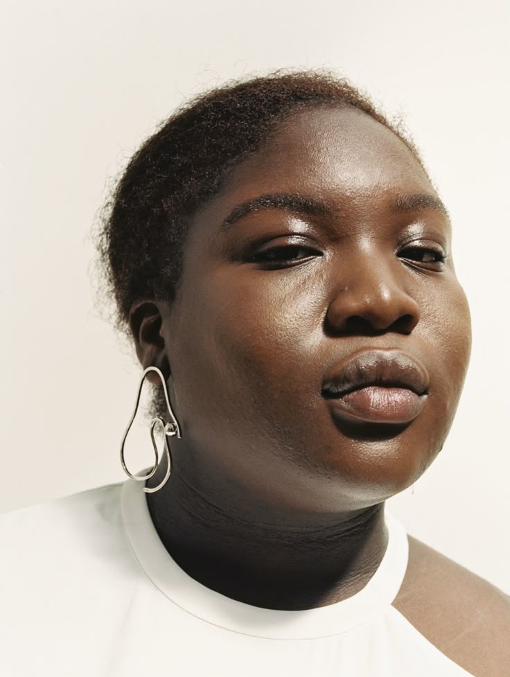 a closeup studio portrait of a darkskinned woman. she is wearing a white shirt and a silver earring of abstract shape. her skin is glossy and she has short hair. the shot focuses on her head and shoulders.