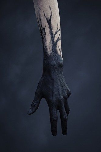 a closeup photo of a person's arm and hand, tattooed with a dark tree. the arm is painted black from the wrist to the elbow, and the hand is painted black with the exception of the fingertips. the photo is taken against a dark blue background.