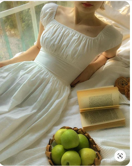 a woman in a white dress is lying on a bed with a book and a basket of green apples. the image is brightly lit, suggesting a daytime setting. the overall aesthetic is soft and romantic.