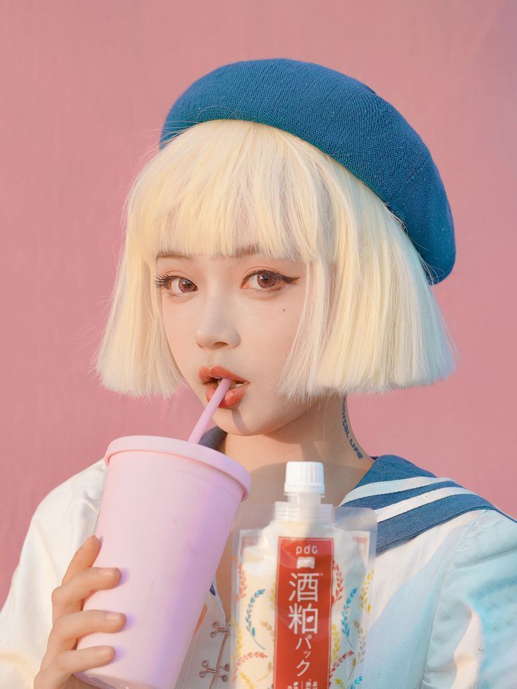 closeup shot of a young woman with blonde bob hair and bangs, wearing a blue beret and sailorcollared shirt. she's holding a pink cup with a straw and a pouch of japanese rice milk drink against a pastel pink background. the overall aesthetic is cute and kawaii. the image is welllit and styled with a focus on pastel colors and soft lighting.