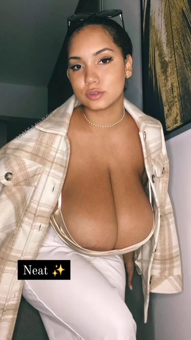 the image shows a closeup portrait of a darkhaired, tanskinned woman with large breasts. she is wearing a beige plaid shirt, white pants and a delicate necklace. she's posed confidently, with sunglasses perched on her head. the setting appears to be indoors. the overall style is sensual and fashionforward, highlighting the woman's curves and attractive features. the picture is sharply focused on the subject.  the word "neat" is superimposed on the bottom left corner.