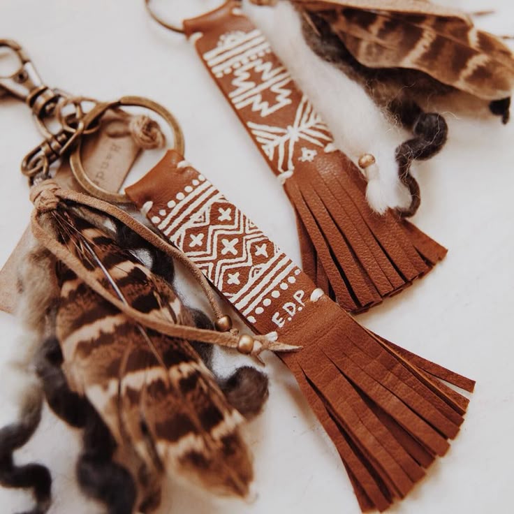 the image shows two leather keychains with feathers and tassels. both keychains have brown leather straps with white painted patterns on them. one keychain has the initials "e.d.p" painted on it. both keychains also have a leather tassel at the bottom, and feathers attached to the keyrings. the feathers are a mix of brown, white, and gray.