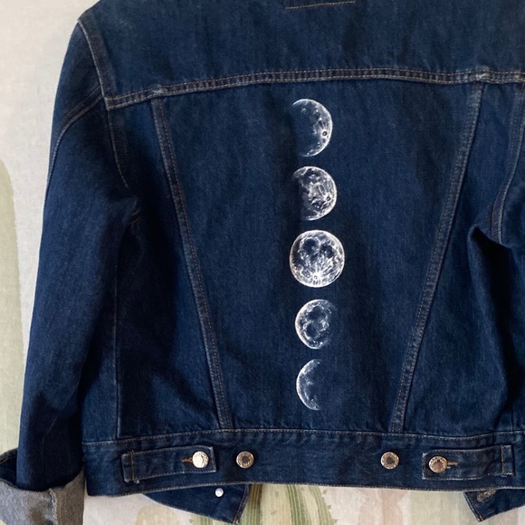 a dark blue denim jacket, neatly buttoned, hangs against a creamcolored wall adorned with a cactus plant. the back of the jacket is embellished with a vertical row of handpainted white moon phases, showcasing the moon's transition from full to crescent.