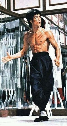 a photo of bruce lee, the famous martial arts master, in a fighting stance. he is wearing black pants and white socks with black shoes. his chest is bare and he is looking intensely at the camera. the photo is taken outside in an urban environment, with a reflection of bruce lee and the background in the window behind him.