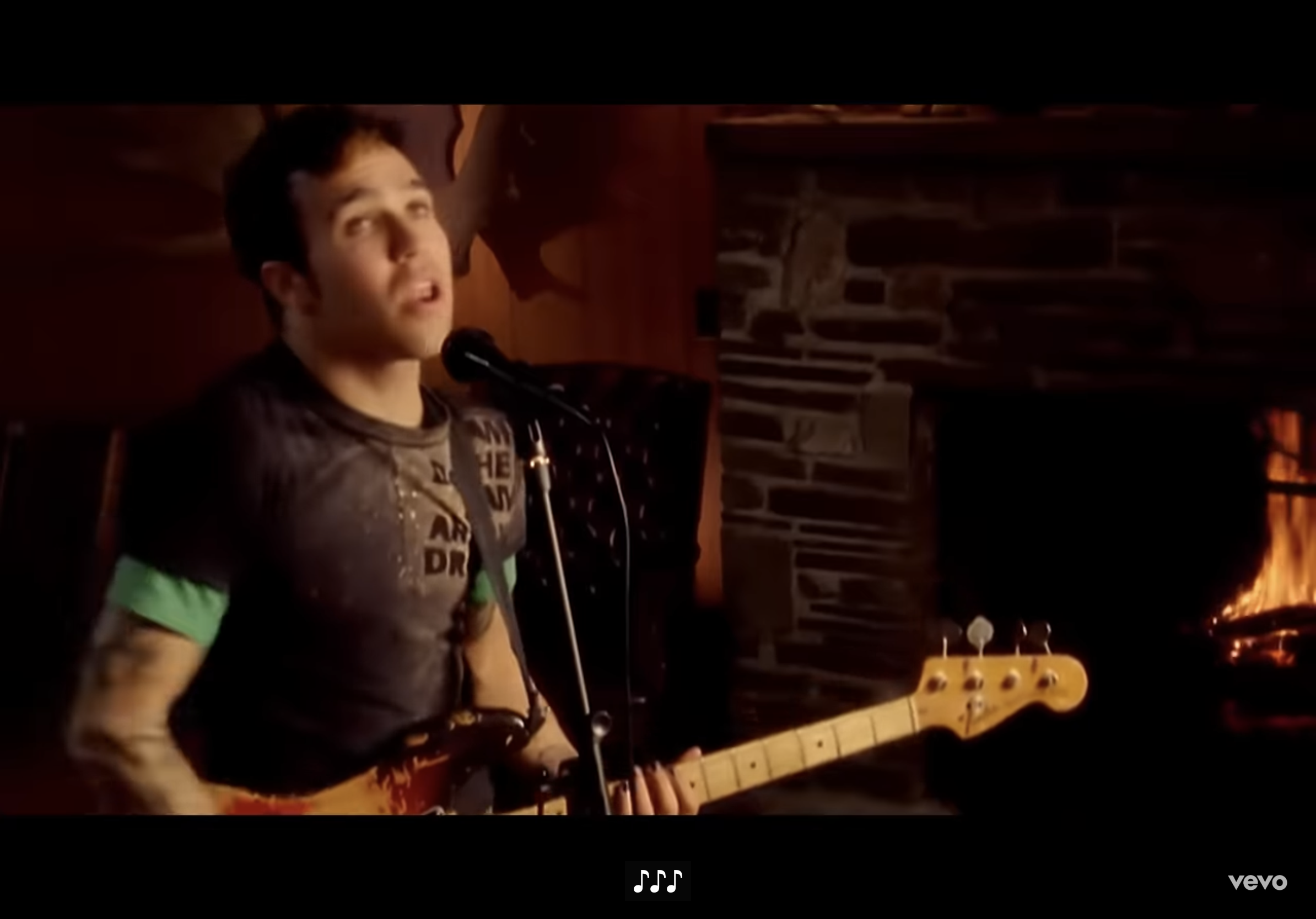 a man with short dark hair and tattoos on his left arm is shown in an eyelevel view, singing and playing a bass guitar. he's wearing a shortsleeved tshirt and is standing in front of a stone fireplace with a roaring fire. the lighting is warm and artificial. the overall ambiance is cozy and intimate.