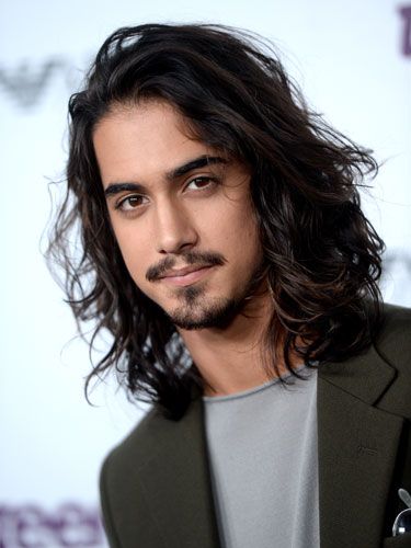 a portrait of avan jogia with long dark hair, a mustache, and a beard. he is wearing a dark green suit jacket over a gray tshirt.