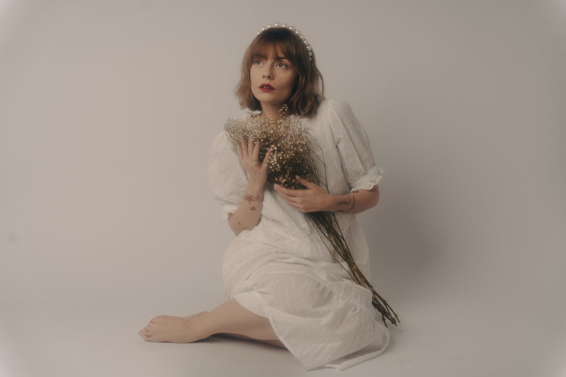 a young woman with short brown hair wearing a white dress and a pearl headband sits on a white background, holding a bouquet of dried baby's breath flowers close to her chest. she is looking upward with a serene expression. the overall style of the image is minimalist and elegant, creating a soft and delicate atmosphere.