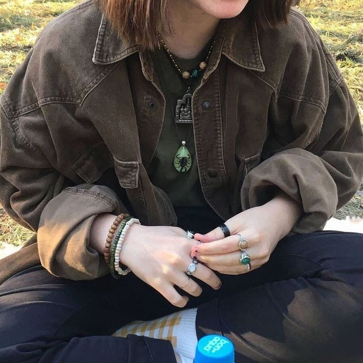a woman wearing a brown corduroy jacket and a green shirt sits on the ground. she is wearing multiple rings and bracelets on her hands. she has a necklace around her neck. the background shows a field with green grass.