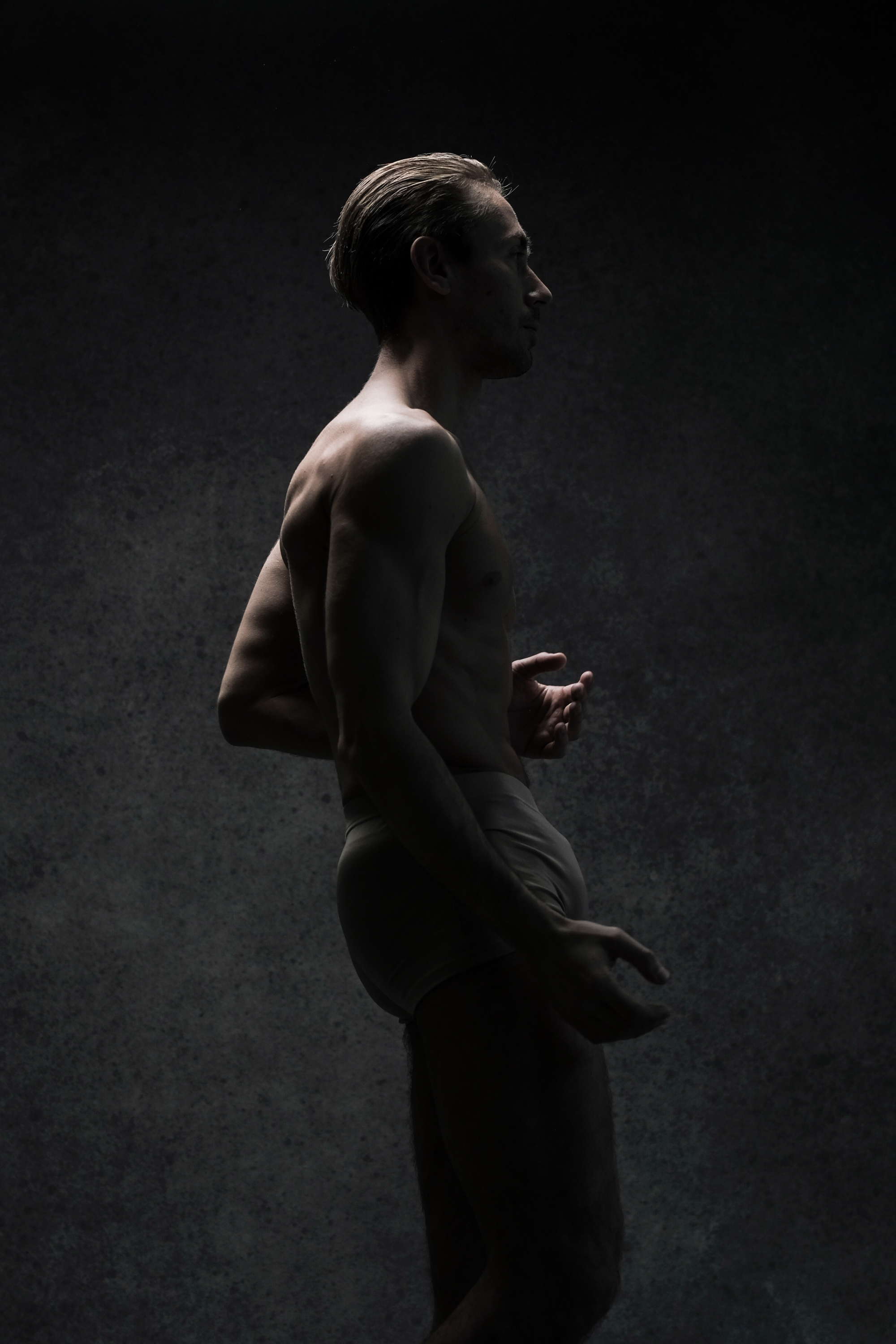 a man in underwear stands in profile against a dark grey background. his muscular body is lit with directional lighting, highlighting his form and the contours of his muscles. his hands are delicately positioned, suggesting a sense of poised movement.