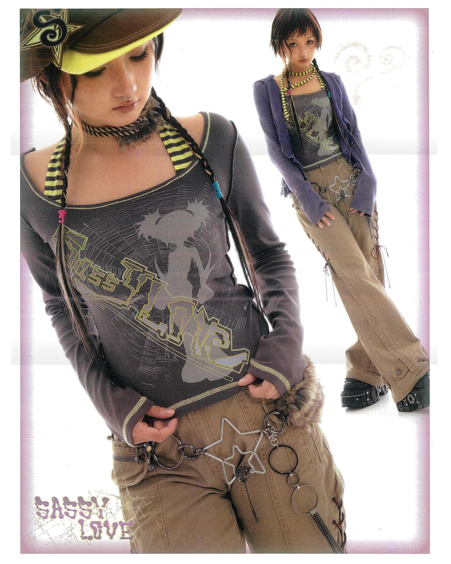 he image showcases two young women styled in a blend of japanese fashion aesthetics, possibly visual kei or a subgenre of lolita fashion with streetwear influences.  both models are wearing distinct yet complementary outfits.  the model in the foreground wears a dark gray longsleeved top with a graphic design, khaki cargo pants, a baseball cap, and an elaborate braided hairstyle accented with colored strings. she also sports a multiring belt featuring a star charm. the other model is dressed in a purple cardigan, a printed top, and khaki pants, complemented by layered necklaces and chunky platform boots. both outfits share similar color palettes and display attention to accessorizing. the overall style is edgy, playful and youthful, indicative of harajuku fashion. the backdrop is simple, allowing the clothing and models to be the focus.  the "sassy love" branding is visible in the bottom left corner.