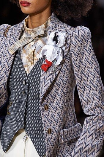 loseup of a female model wearing a sophisticated autumn/winter outfit. the ensemble features a beige and navy herringbone tweed blazer with puff sleeves, layered over a matching waistcoat and a pale yellow plaid shirt. a large, eyecatching brooch with white and red flower details adorns the jacket. the model's hair is styled in a voluminous afro. the overall impression is one of high fashion and refined elegance. the image focuses on the intricate details of the clothing and its textures.