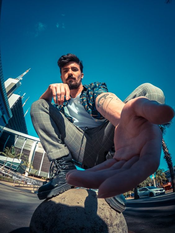 a stylish young man with a beard and tattoos is taking a selfie with a fisheye lens, crouching on a rock in front of modern buildings in a city. the image is vibrant and dynamic, with a strong perspective created by the lens. the man is wearing trendy clothes and his pose exudes confidence and urban style. the bright sunlight creates sharp contrasts and highlights the modern architecture in the background.