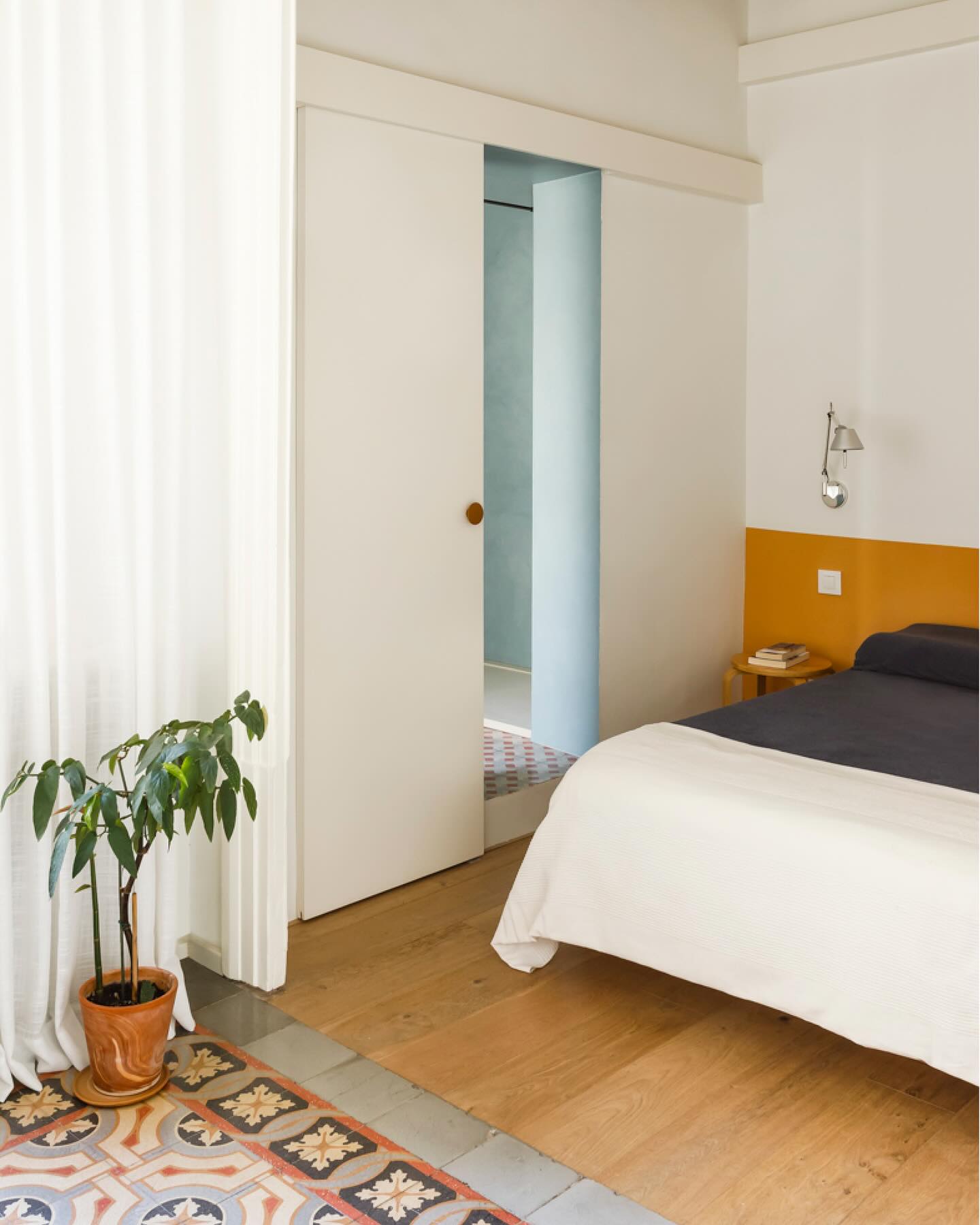 the image shows a bedroom interior. a bed with a white comforter and dark blue sheets is partially visible on the right side of the frame. the headboard is a bright yellow color. a nightstand is located beside the bed with a small lamp and book on it. a wall light is mounted on the wall above the nightstand. a potted plant sits on the floor in the foreground, next to a white curtain. the floor is made of wood, except for a tiled section near the plant. a sliding door is slightly open, revealing a glimpse into another room with blue walls and tiled floor.