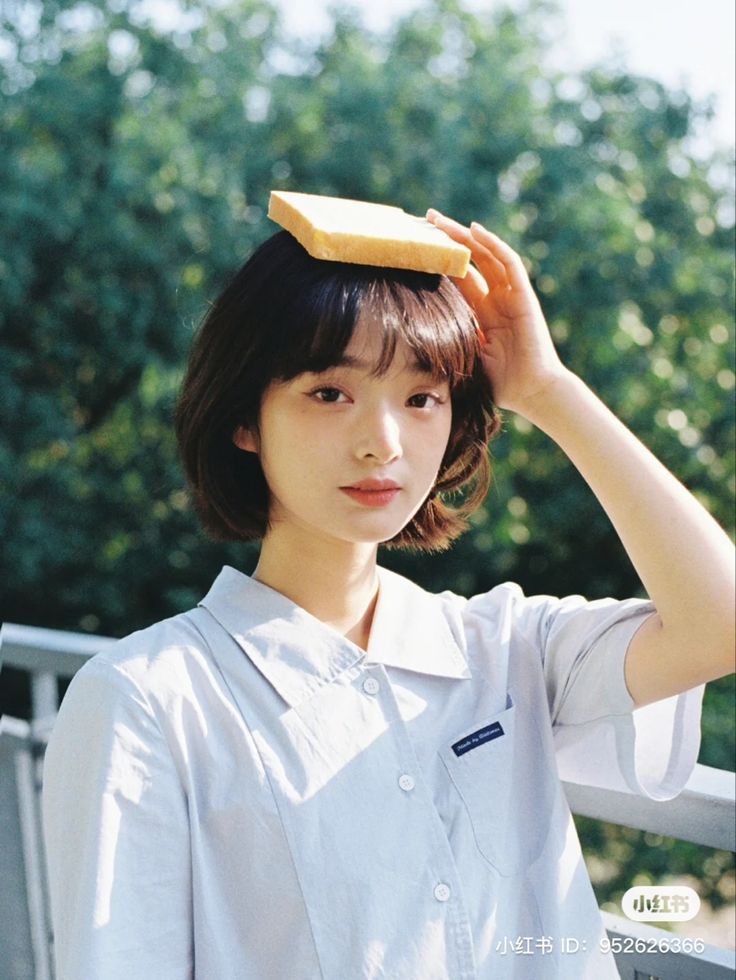 a young woman with short brown hair, wearing a white buttondown shirt, stands outdoors in a sunny summery setting with a slice of bread on her head. she looks playful and cute, smiling softly at the camera. the image is bright and soft, capturing a sweet and innocent moment.
