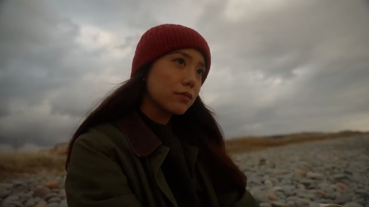 a young woman with long black hair wearing a red beanie and a green jacket sits on a rocky beach. the sky is overcast with dark grey clouds, creating a moody and atmospheric setting. the woman has a contemplative expression on her face as she looks out at the distance.