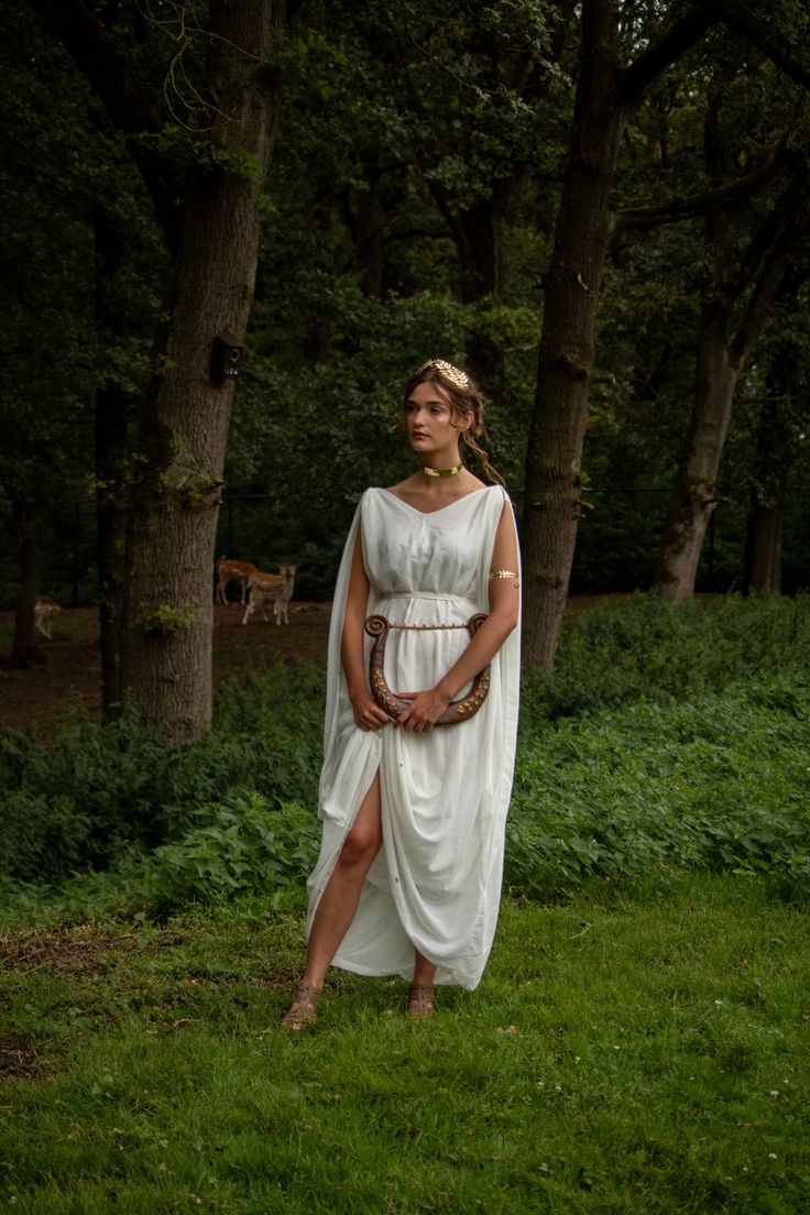 a full shot of a woman in a white ancient greekstyle dress standing in a forest. she is holding a lyre and wears gold jewelry.  the scene is serene and peaceful, suggesting a classical or mythological setting.  the background is lush greenery and tall trees.