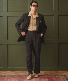 a man is standing in front of a green wall wearing a black suit and white shirt with a pattern. he has on brown shoes, sunglasses, and a patterned tie. he is posing with his hands in his pockets.
