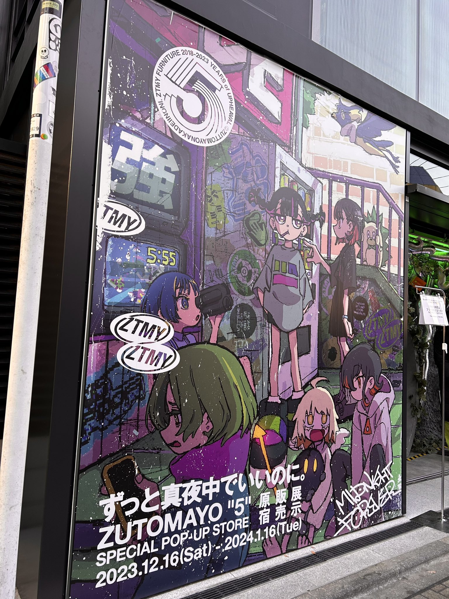 a large, colorful poster advertising a zutomayo exhibition and popup store in japan. the poster features animestyle characters and japanese text. it's plastered on the glass exterior wall of the store, illuminated by overhead lights.
