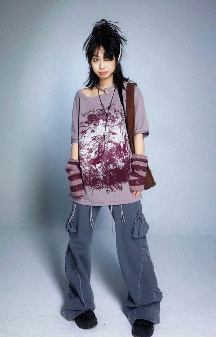 a fullbody shot of a young woman posing in a studio against a light gray background.  she's wearing a loosefitting,  purple graphic tshirt with a natureinspired design, baggy gray cargo pants, knit wrist warmers, and a brown handbag slung over her shoulder.  she also has on a necklace and headphones around her neck. her hair is styled with bangs and falls to her shoulders. she's looking toward the camera with a slightly playful expression. the overall style is relaxed and trendy, leaning towards a slightly grunge or alternative aesthetic.