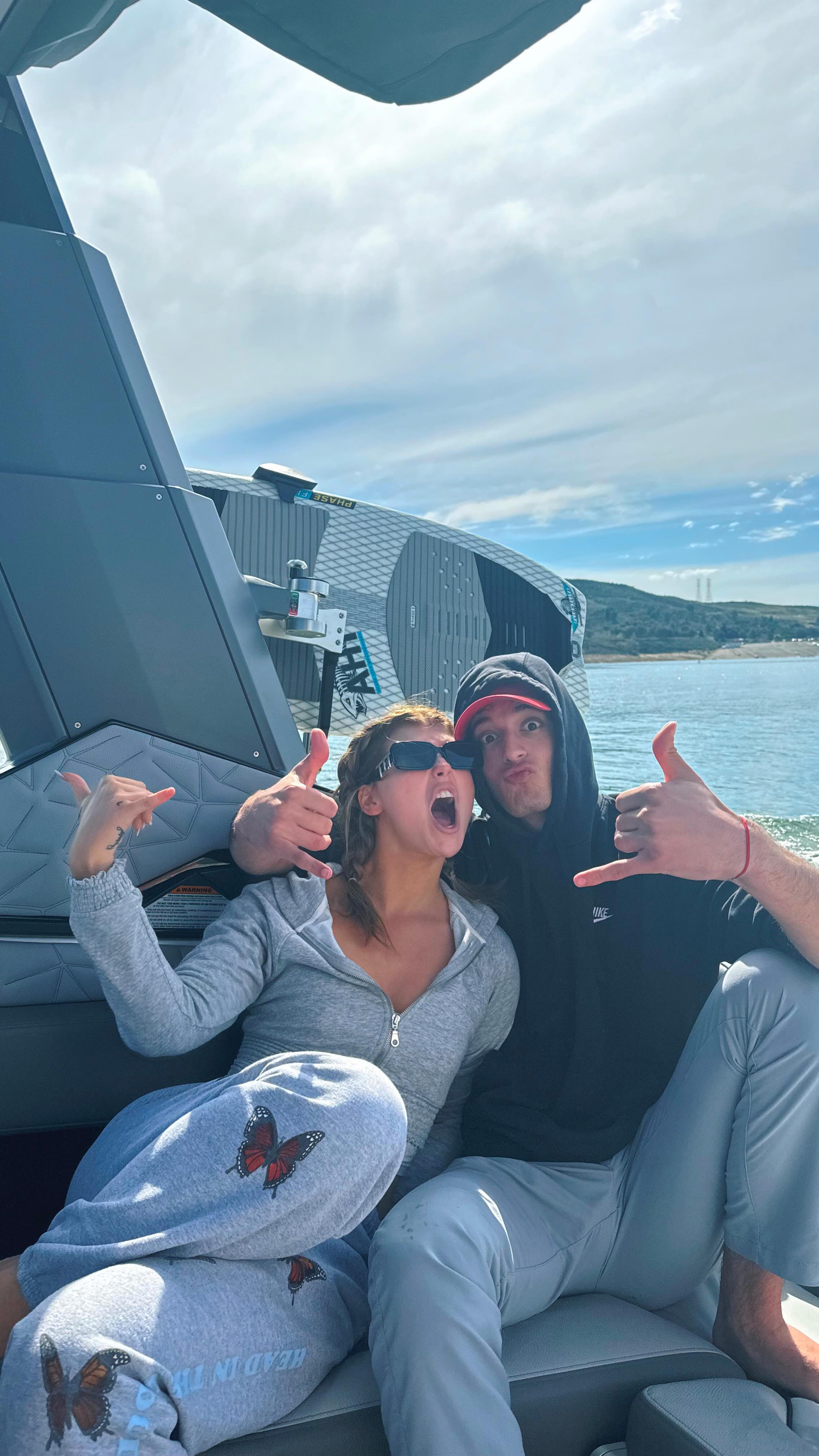 a woman and a man are sitting together on a boat on a lake. the woman is wearing sunglasses, a grey zipup hoodie, and grey sweatpants with butterflies on them. she has her mouth open and is making a "shaka" hand gesture with her left hand. the man is wearing a dark hoodie, a red baseball cap and grey sweatpants. he also doing the "shaka" sign with his right hand and resting it on the woman's shoulder. they are smiling and the background shows a body of water with trees on the shore.