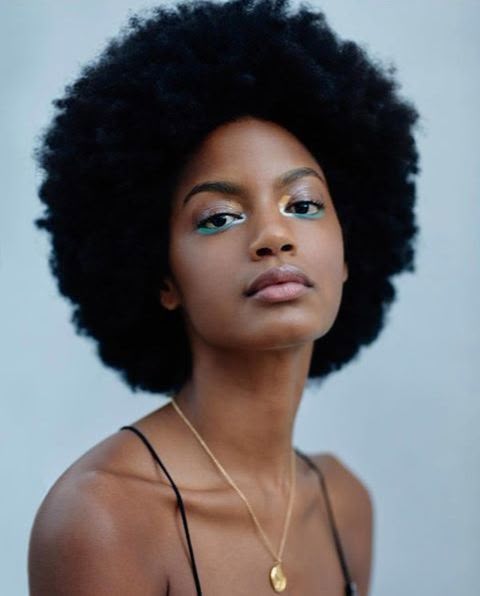 a closeup portrait of a beautiful woman with a large afro. she is wearing makeup, including gold and blue eyeshadow and nude lipstick. she is also wearing a gold chain necklace with a round pendant. she is wearing a black spaghetti strap top. she is looking directly at the camera with a serious expression. the background is a light blue.