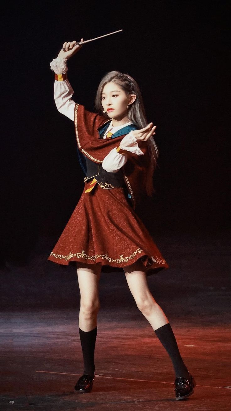a young asian woman, possibly a singer or idol, is captured midperformance on a dark stage. she's dressed in an elegant red dress with gold detailing, resembling a conductor's outfit. she holds a baton, suggesting she might be conducting a musical piece. her posture is graceful, and her expression is focused and poised. the overall mood is one of elegance and performance. the dark background highlights the woman and her attire.