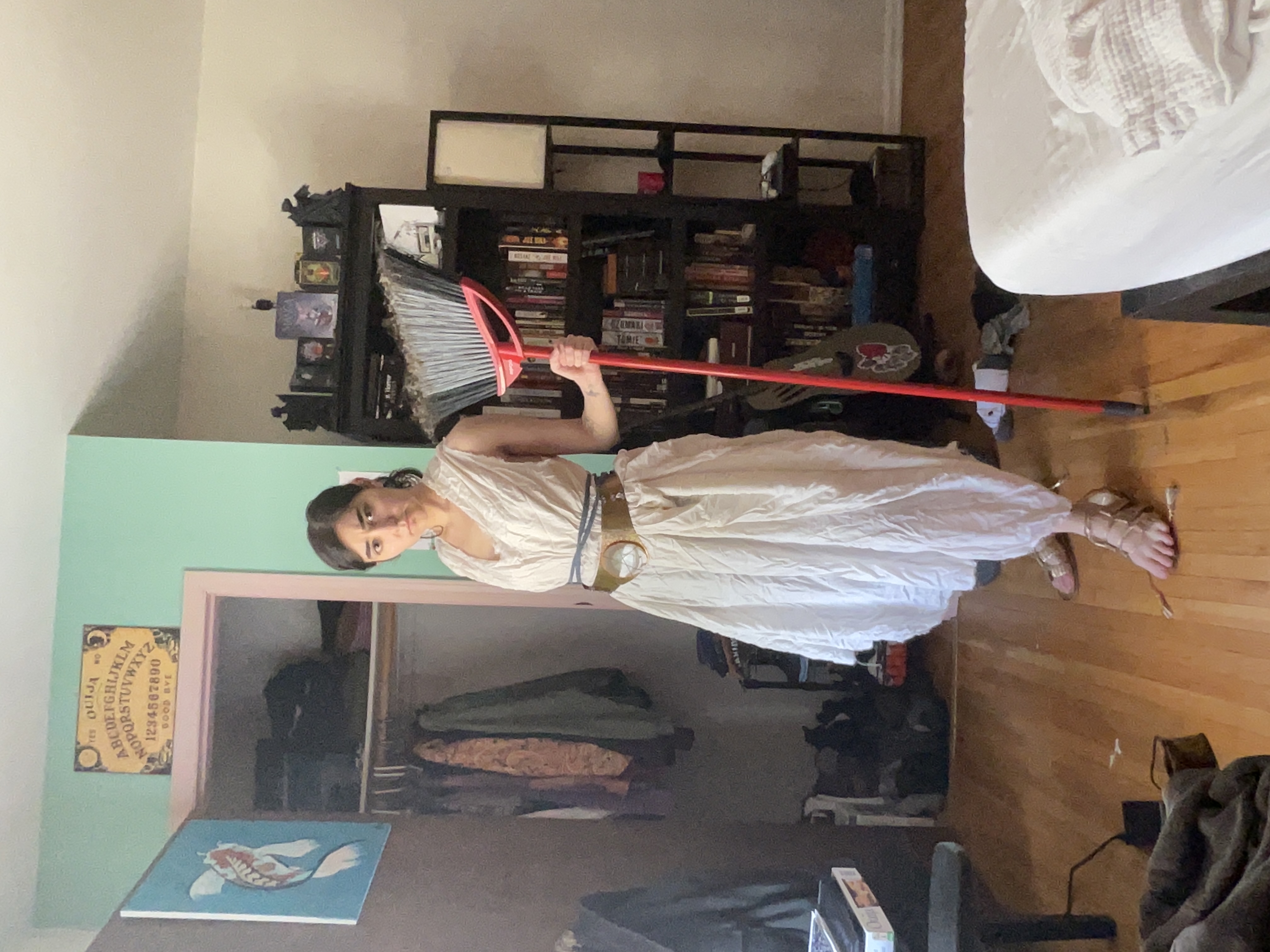 a woman in a white dress and sandals holds a red broom in a bedroom. the room has a bookshelf, closet, and bed. a framed ouija board image hangs above the closet door.