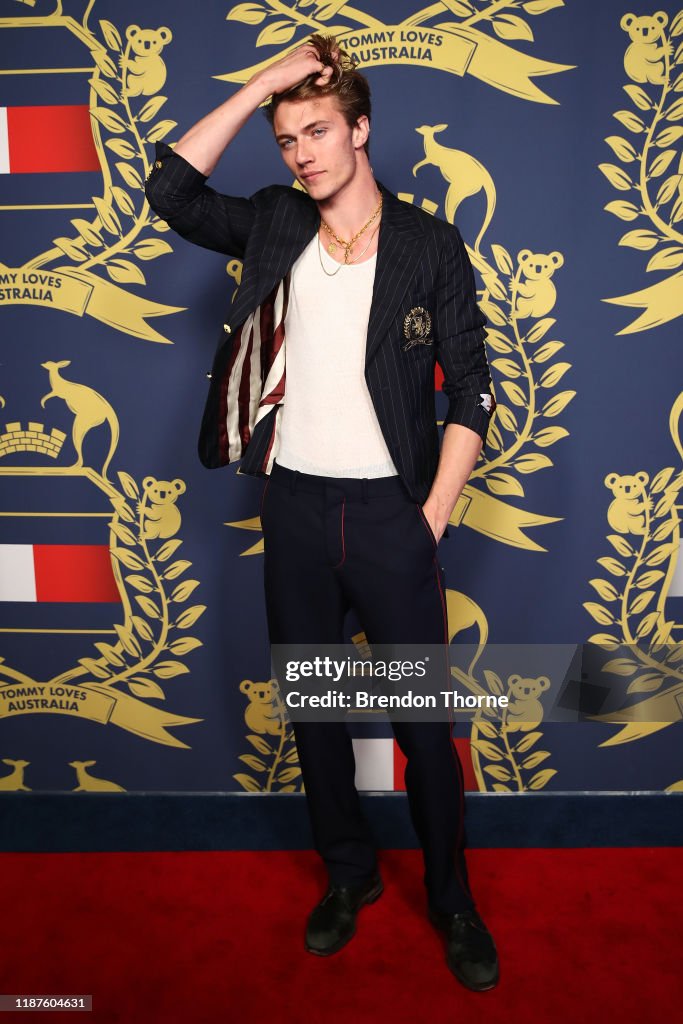 a man wearing a blazer, trousers, and dress shoes poses on a red carpet in front of a "tommy loves australia" backdrop. he is wearing a white tshirt and a gold necklace underneath the blazer. he has his hand in his hair.
