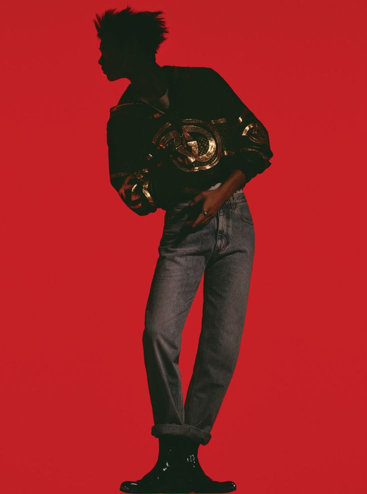 a man in a black and gold patterned jacket poses against a red background. he is wearing blue jeans, black boots, and has a slight smile on his face. the image is shot in a studio setting with a minimalist background. the man's silhouette is emphasized by the bright red background.