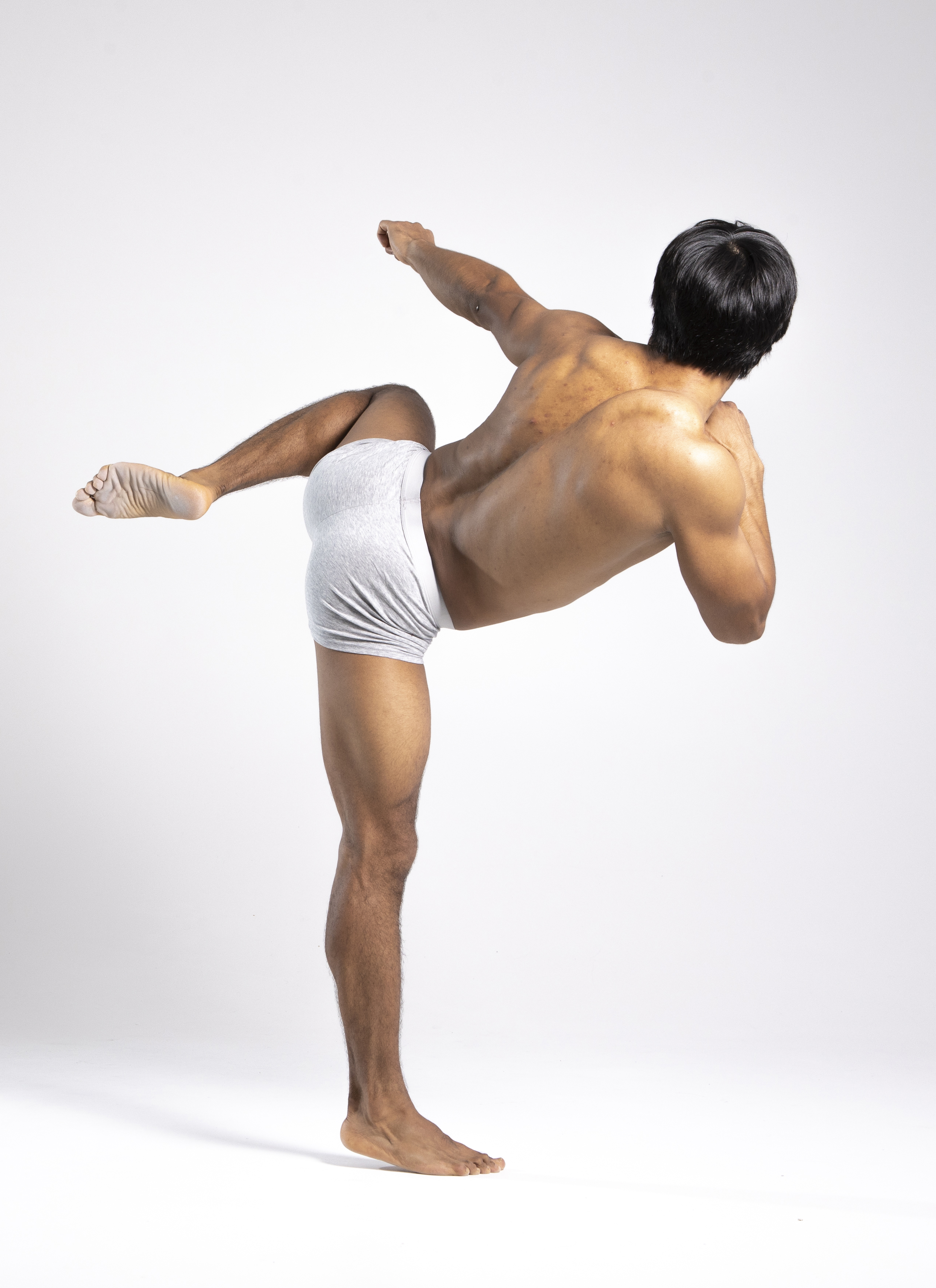 a muscular man is standing in a martial arts pose, kicking his leg out to the side. he is wearing grey underwear and has his back to the camera. his arm is extended to the side and he is looking to the right. the background is white.