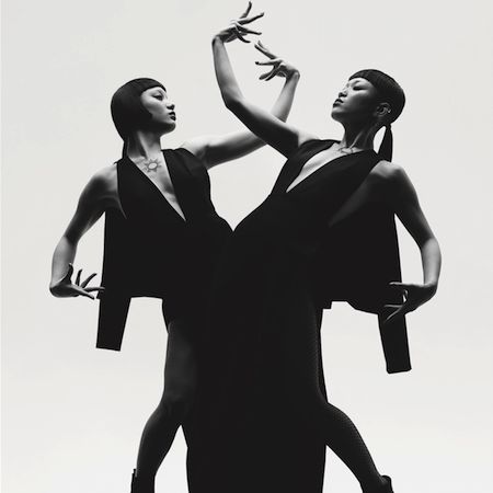 two women dressed in black are posed in a studio, they appear to be dancing. the woman in the front is holding her arms above her head, while the woman behind her has one arm wrapped around her waist.  their hair is styled in a simple fashion, and their clothes are sleek and modern. their postures are graceful and elegant. they appear to be contemporary dancers.