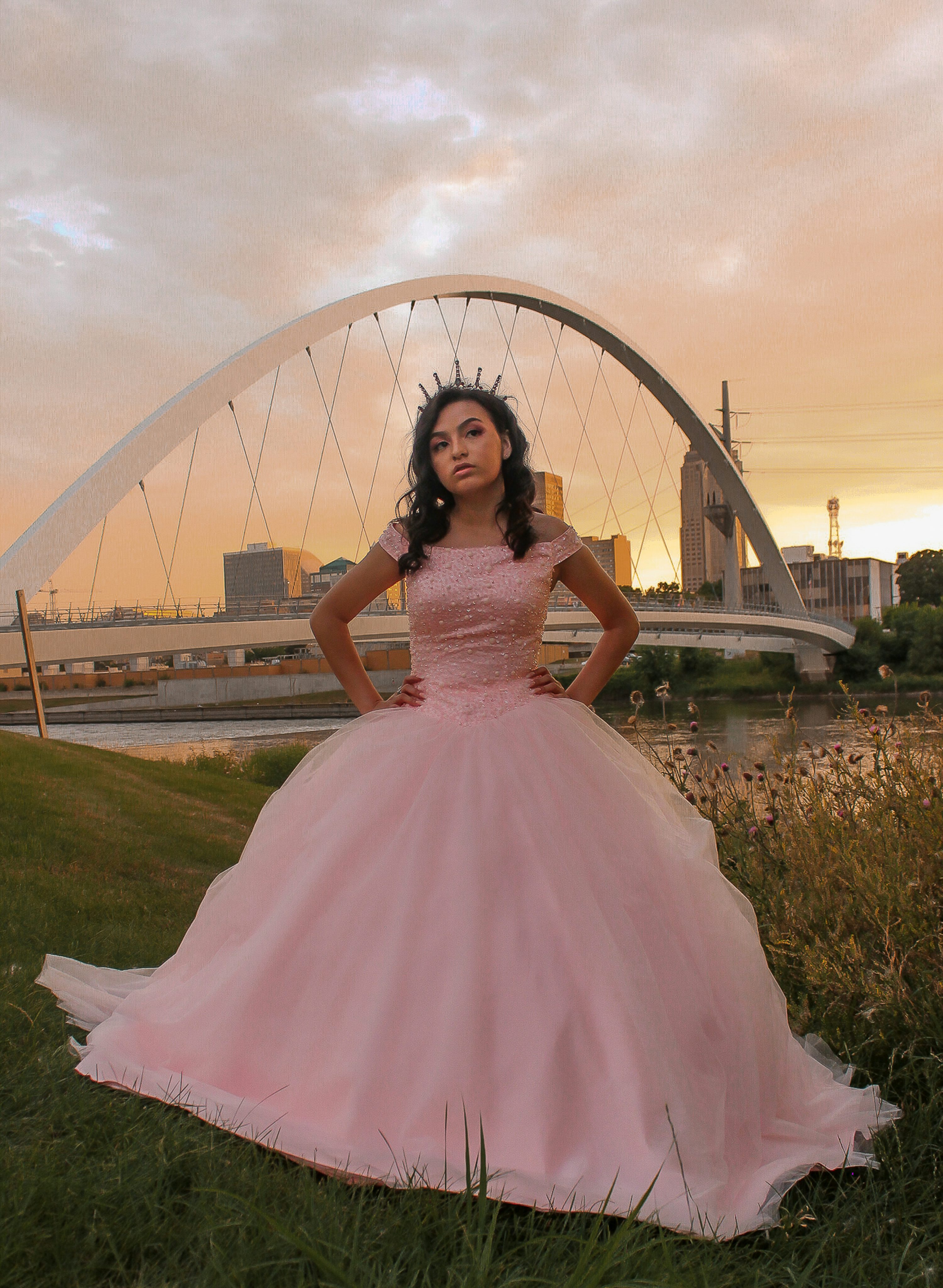 a young latina woman, possibly celebrating her quinceañera, stands confidently in a beautiful, flowing pink ball gown. she wears a tiara and stands with her hands on her hips. the setting is outdoors, with a modern, arched bridge and cityscape in the background, as the sun sets, casting a warm, golden light. the scene is elegant and evokes a sense of celebration and joy. the overall feel is one of grace and sophistication.