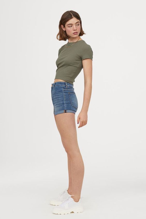 a young woman stands against a white background wearing a green tshirt, blue denim shorts, and white sneakers. she has brown hair and is posing with her hand on her thigh. the image is brightly lit and taken from an eyelevel perspective.