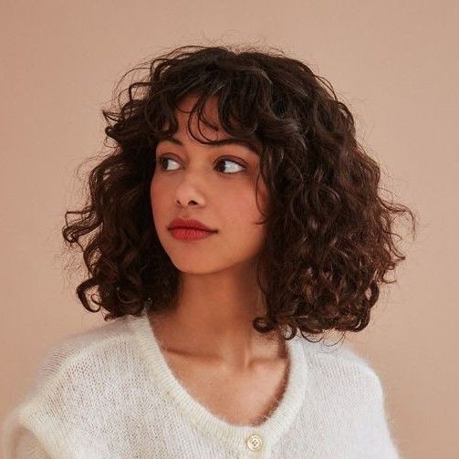 a headshot portrait of a woman with brunette, curly, shoulderlength hair and bangs. she is wearing a white cardigan and red lipstick, looking to the left of the frame. the background is a solid, neutral color.