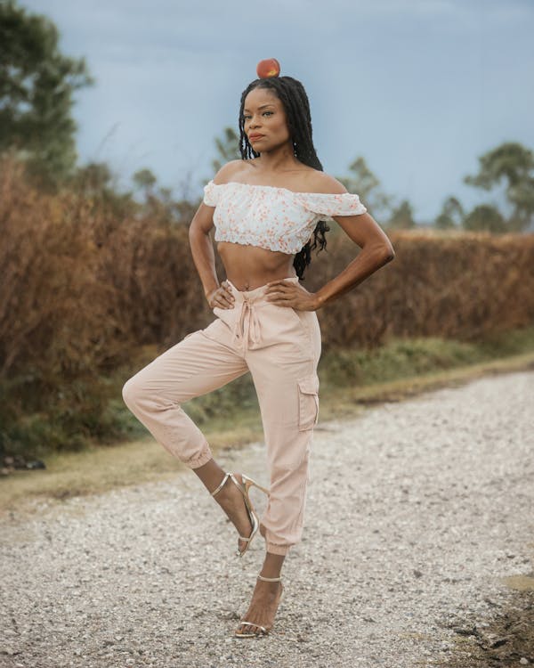 a fullbody shot of a stylish woman posing outdoors. she's wearing a blush pink crop top and cargo pants, and gold heels. a peach is balanced on her head. she's standing on a gravel road with a natural background of tall grasses and trees. the overall mood is confident and fashionable.