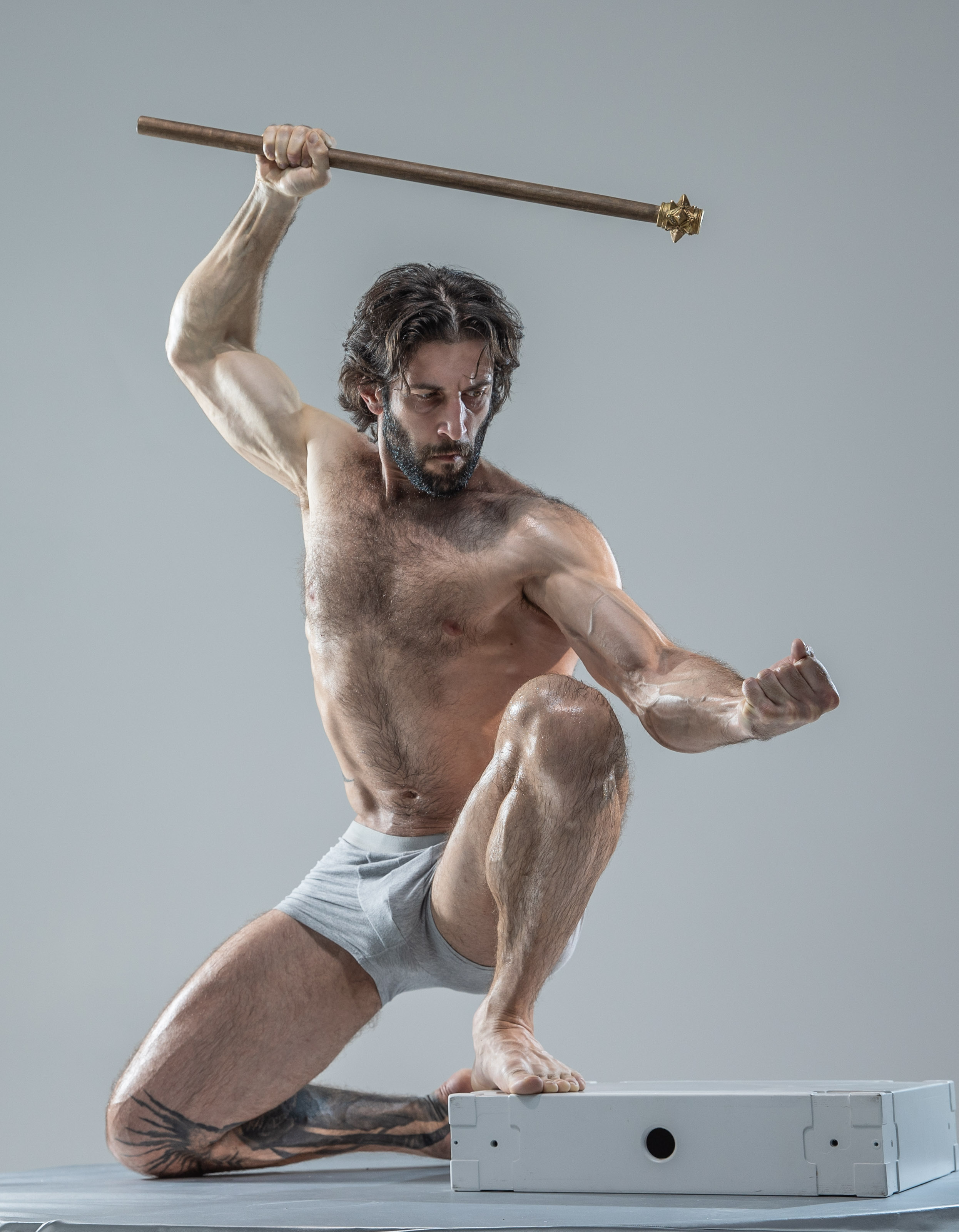 a muscular, bearded man with a determined expression lunges forward in a dynamic pose. he grips a wooden staff with a brass end, suggesting a warrior or fighter. he wears only white underwear, emphasizing his toned physique. the studio setting features a plain grey background and soft, even lighting. the low angle of the shot adds to the sense of power and aggression.