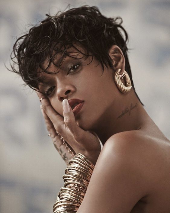 a closeup portrait of a woman with short, wet, curly hair. she is wearing gold earrings and bangles and has a tattoo on her neck. she is looking at the camera with a sensual expression. her lips are painted red and her skin is bare and glowing.