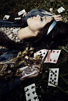 a young woman lies peacefully among flowers and playing cards in a dark and mysterious forest setting. her face is covered in a gentle shadow and she wears a headband. the image has a whimsical feel, reminiscent of a fairytale.