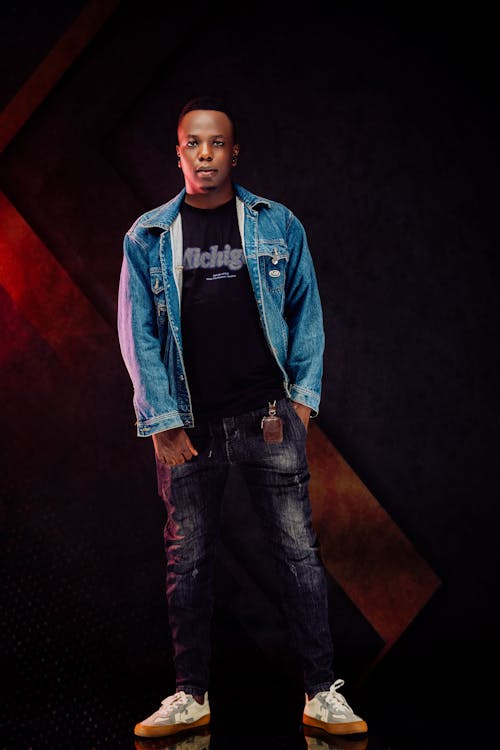 a full shot of a young black man standing confidently against a dark, moody background. he is wearing a denim jacket over a black tshirt, dark wash jeans, and offwhite sneakers. his expression is serious, and his pose is relaxed yet stylish. the background features a diagonal reddishbrown element against a black backdrop, adding a dramatic effect to the image.  the lighting is focused on the man, highlighting his features and clothing.