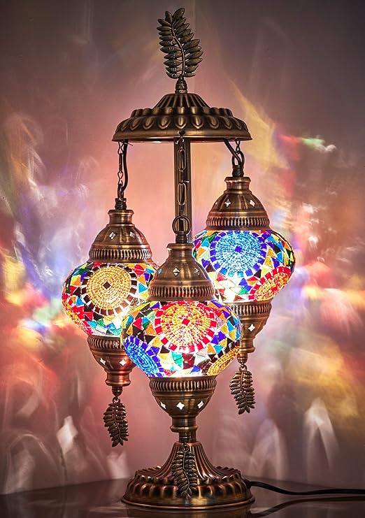 the image shows a decorative turkish mosaic lamp with three hanging lamps. the lamps are made of colorful glass pieces, creating a mosaic pattern when lit. the base and frame are bronze, adding to the ornamental look. the lamp is illuminated, casting warm light and colorful shadows on the surrounding surface. it is placed on a table, suggesting a home decor or interior design setting.
