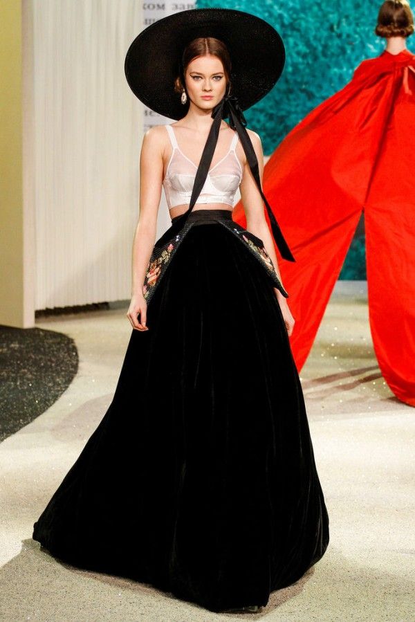 a fashion model walks the runway in a black velvet dress with floral embroidery. she is wearing a white bra underneath and a black hat with a ribbon tied around her neck. the model is walking towards the camera and her dress is flowing behind her. the runway is covered in glitter. another model is visible in the background wearing a red dress.