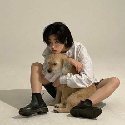 a young woman with dark hair and a white sweater is sitting on the floor, her legs crossed. she is holding a small beige dog in her arms. the woman is wearing black boots and has a casual and relaxed appearance. she is looking down at the dog with a gentle expression. the background is a white studio wall.  this photo captures a moment of companionship between a woman and her pet dog, and it showcases a natural and unpretentious style.